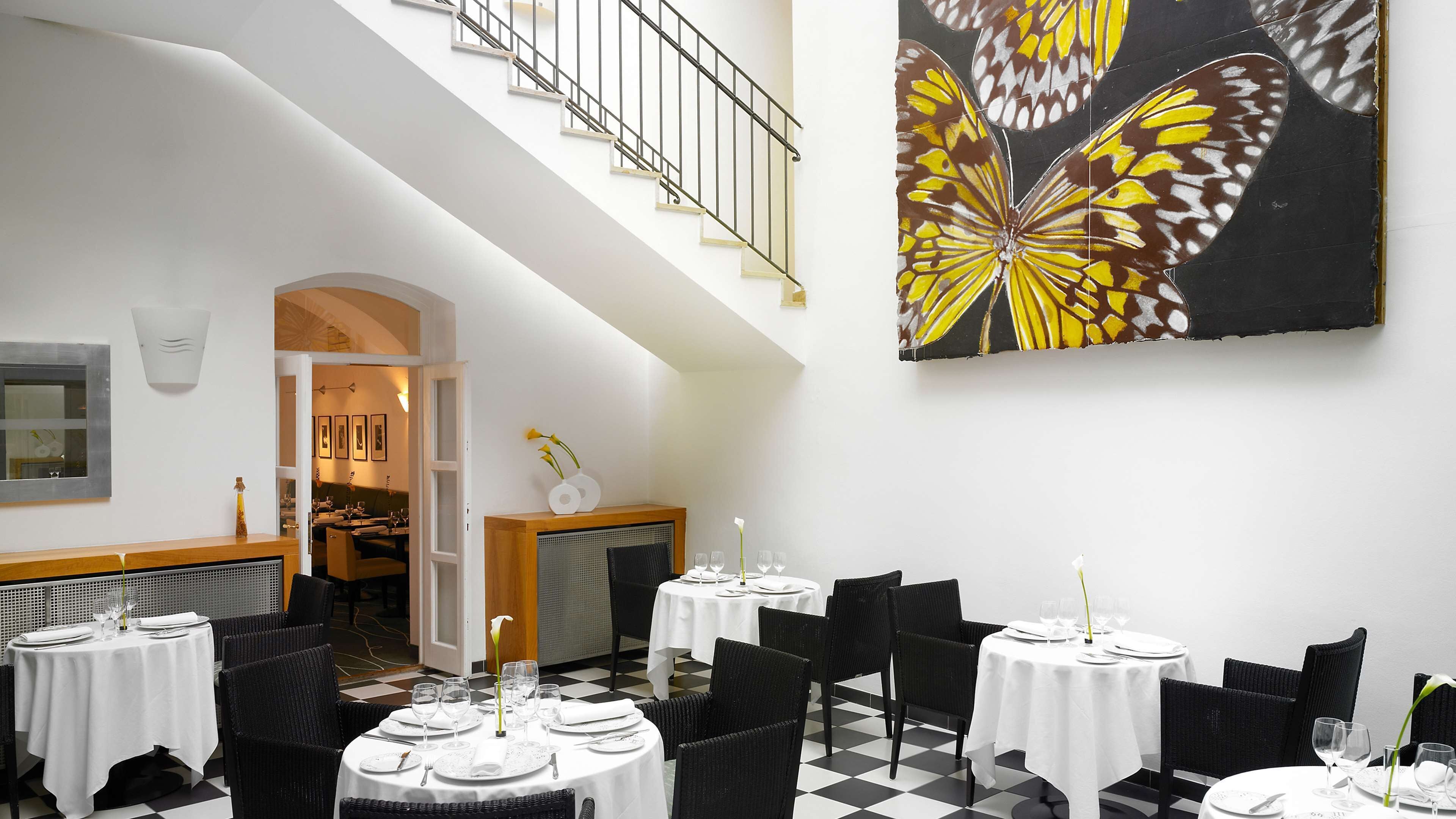 Une salle à manger avec des tables dressées pour le service, un escalier et un grand tableau représentant un papillon accroché au mur.
