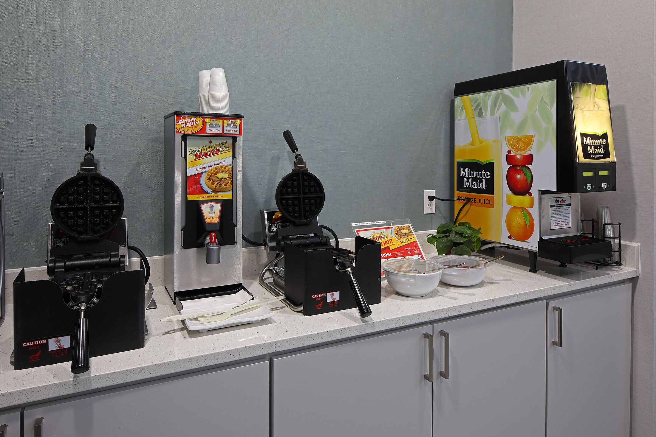 A breakfast bar with waffle makers, a coffee machine, and a juice dispenser.