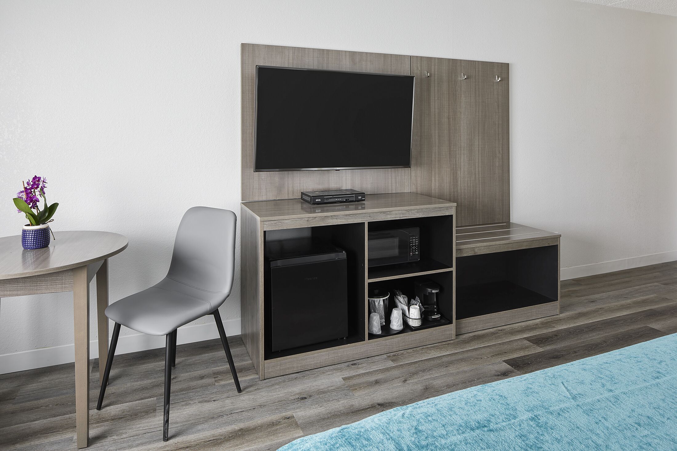 Habitación moderna con televisor de pantalla plana montado en una pared de madera, un refrigerador negro pequeño, microondas y una zona de estar con una silla gris y una mesa con un jarrón de flores.