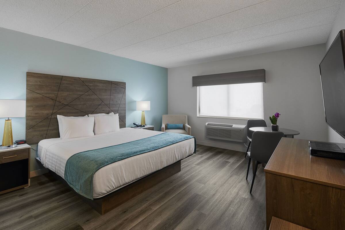 A hotel room with a large bed, bedside tables, a desk, a chair, a TV, and a window with blinds.