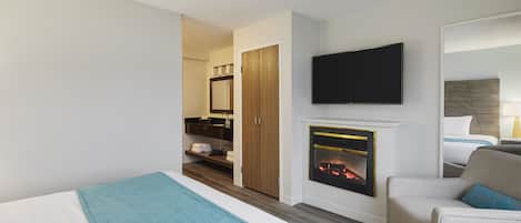A hotel room with a bed, a TV mounted on the wall, a fireplace, and a seating area with a chair and a sofa.