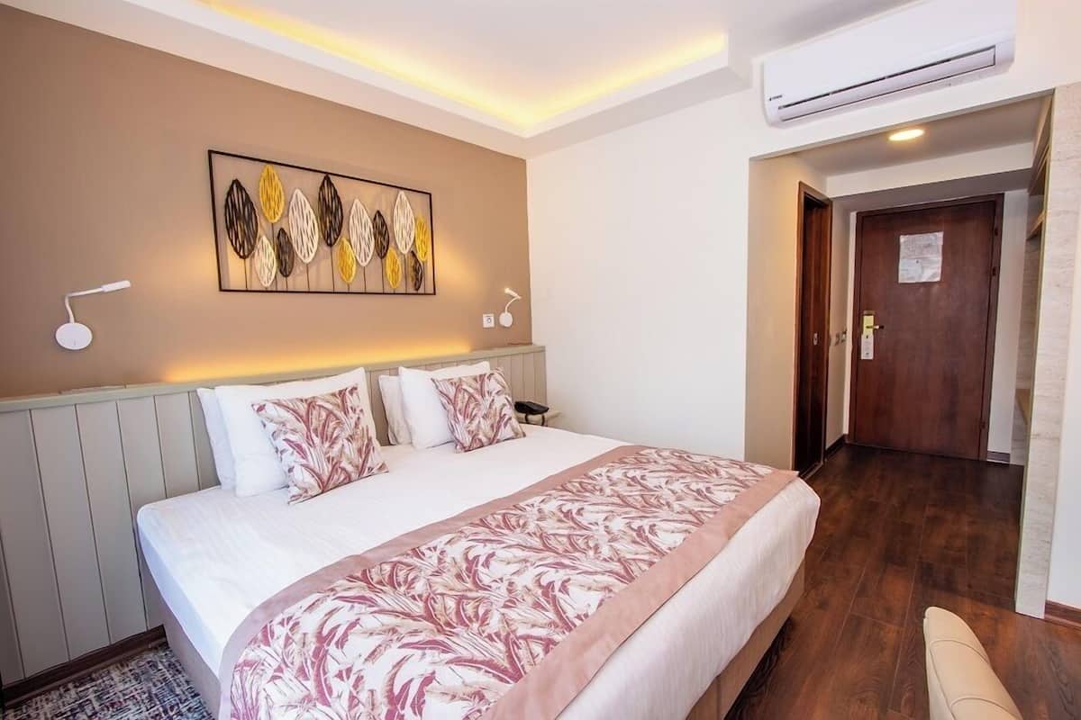 A hotel room with a bed, a wall-mounted artwork, an air conditioning unit, a bedside lamp, and a door leading to another room.