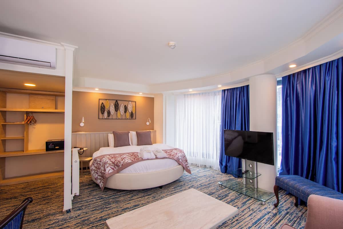 A hotel room with a round bed, a flat-screen TV, a sofa, a glass coffee table, and a wall-mounted shelf.