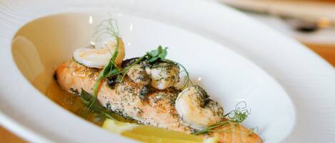 Un plato blanco con un plato de mariscos, una copa de vino blanco y un jarrón con flores amarillas sobre una mesa de madera.