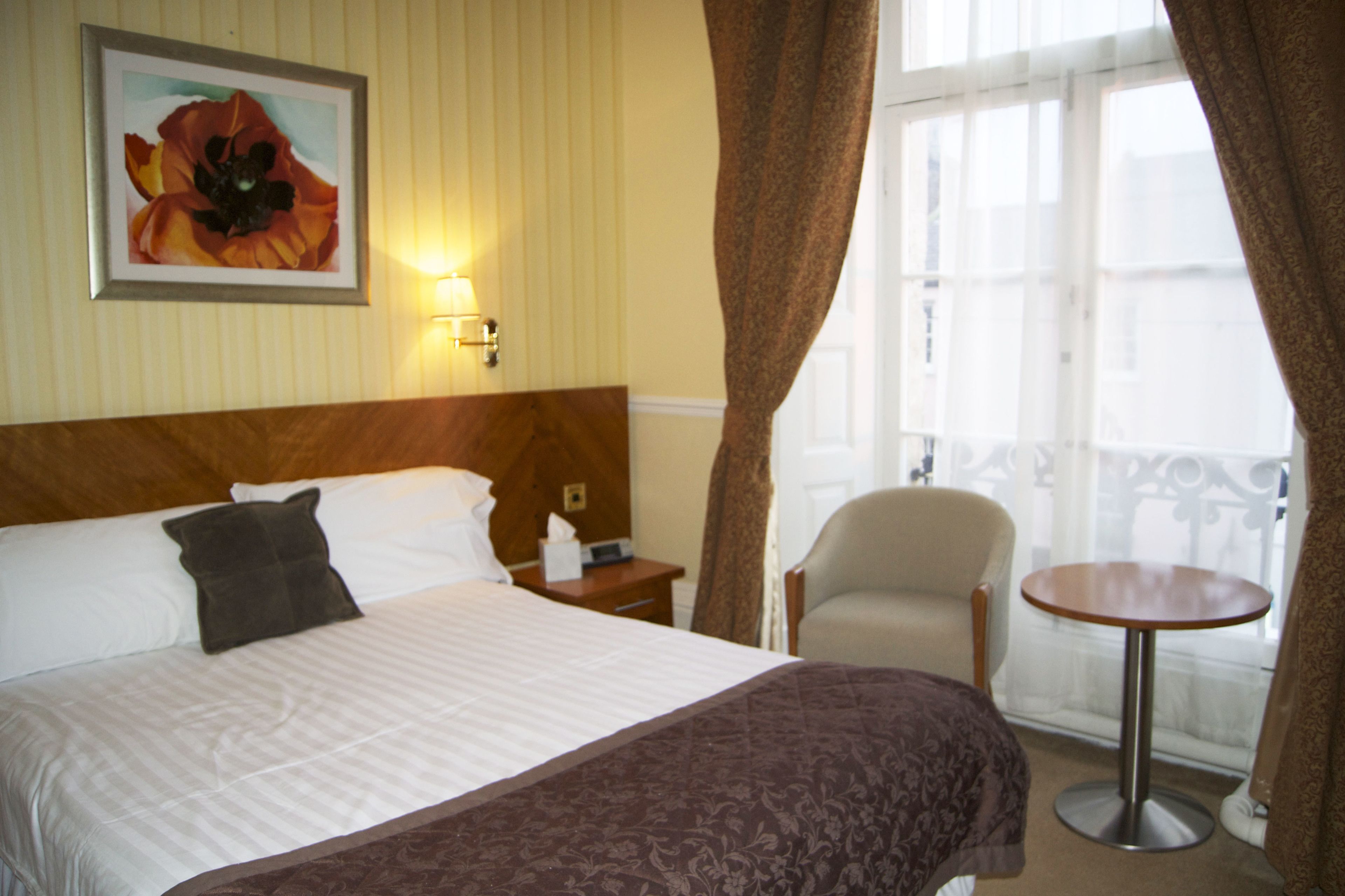 A hotel room with a bed, a chair, a small table, a painting, and a window with curtains.