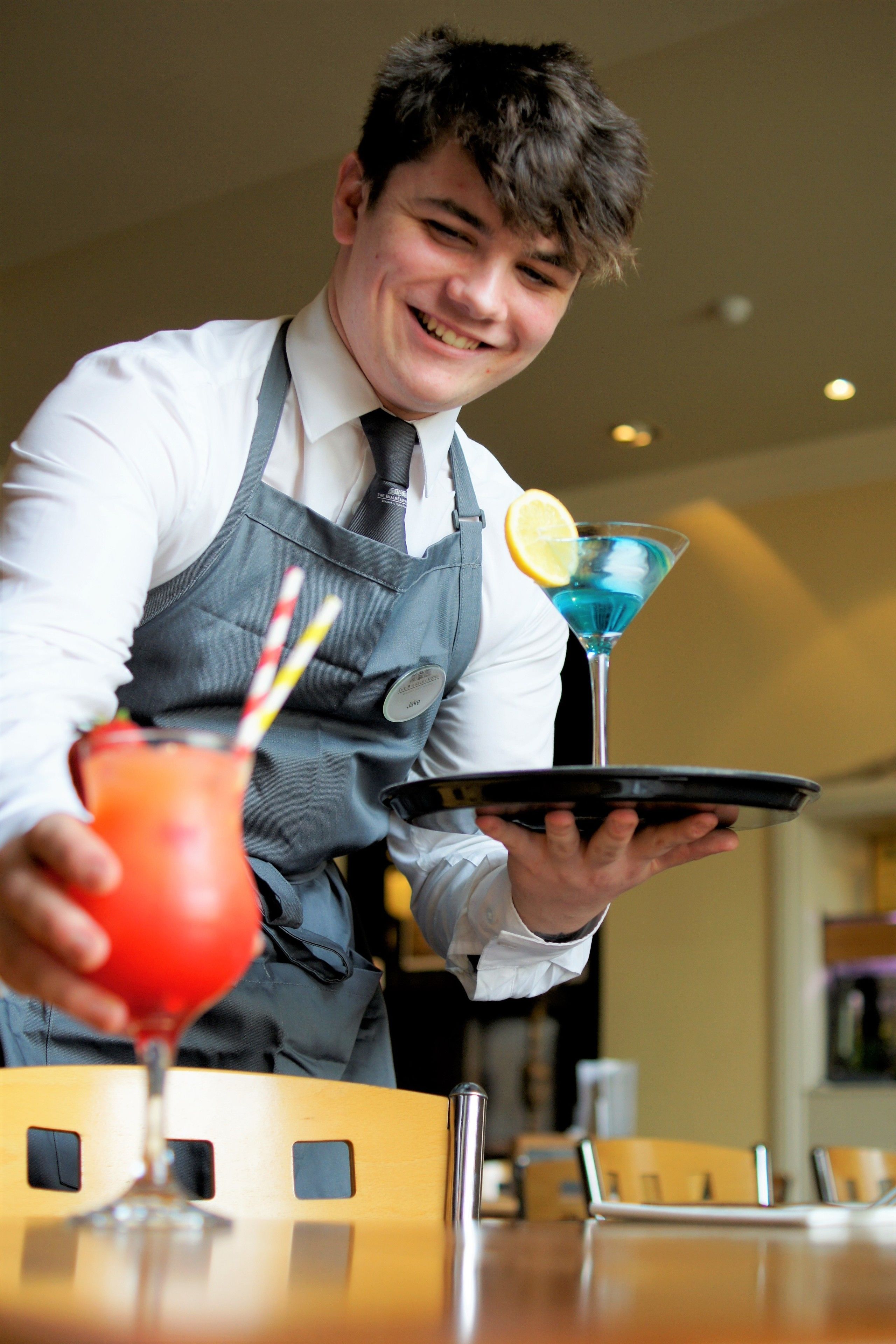 A person in a white shirt and gray apron holding a tray with a blue cocktail and a red drink with a straw.