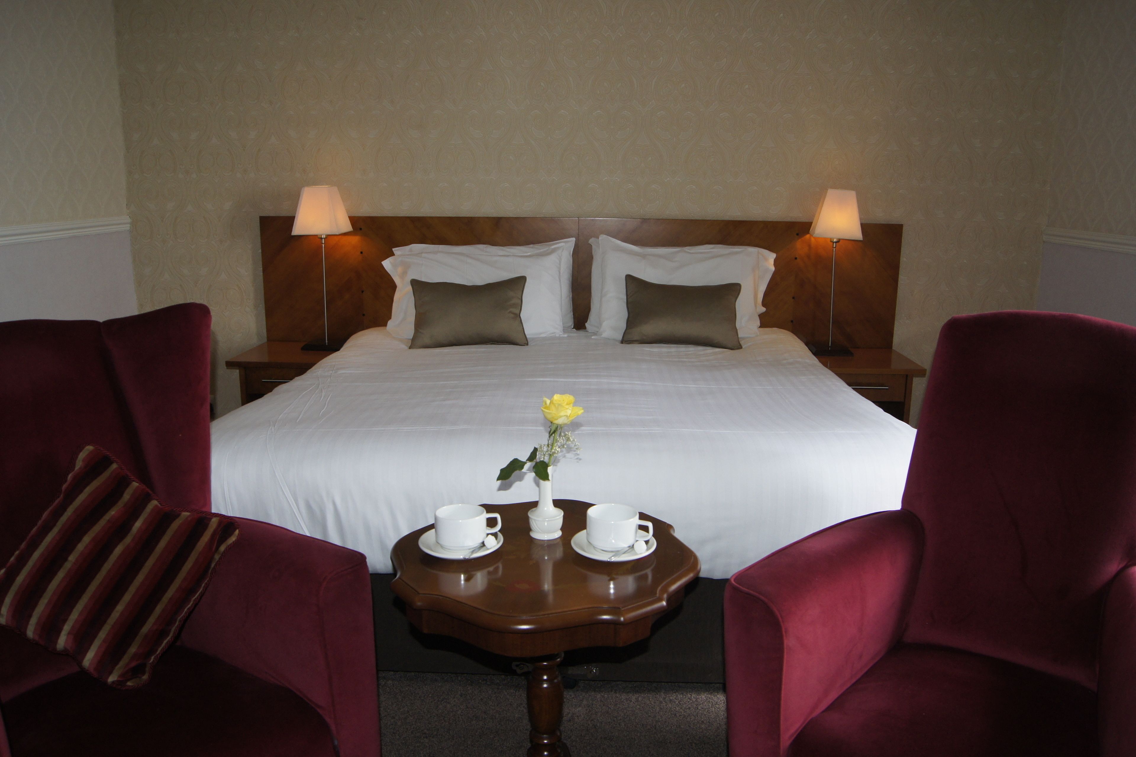 Habitación de hotel con una cama grande, dos lámparas de noche, una mesita con dos tazas y un florero con una flor, y dos sillones rojos.