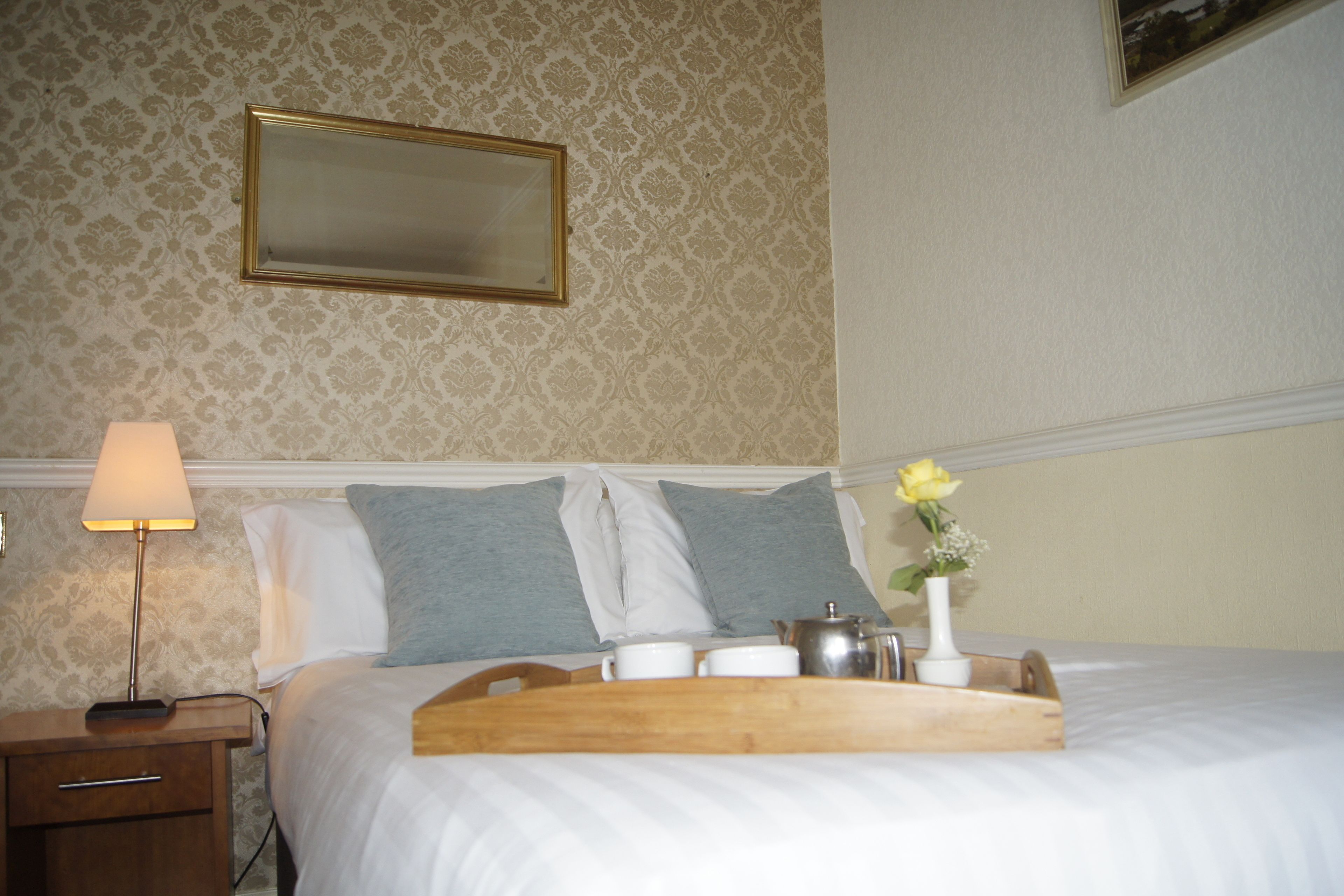 A neatly made bed with pillows, a bedside table with a lamp, a tray with cups and a vase of flowers, and a mirror on the wall.