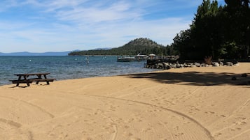 Una playa arenosa con una mesa de picnic, agua tranquila, barcos y una colina boscosa al fondo.