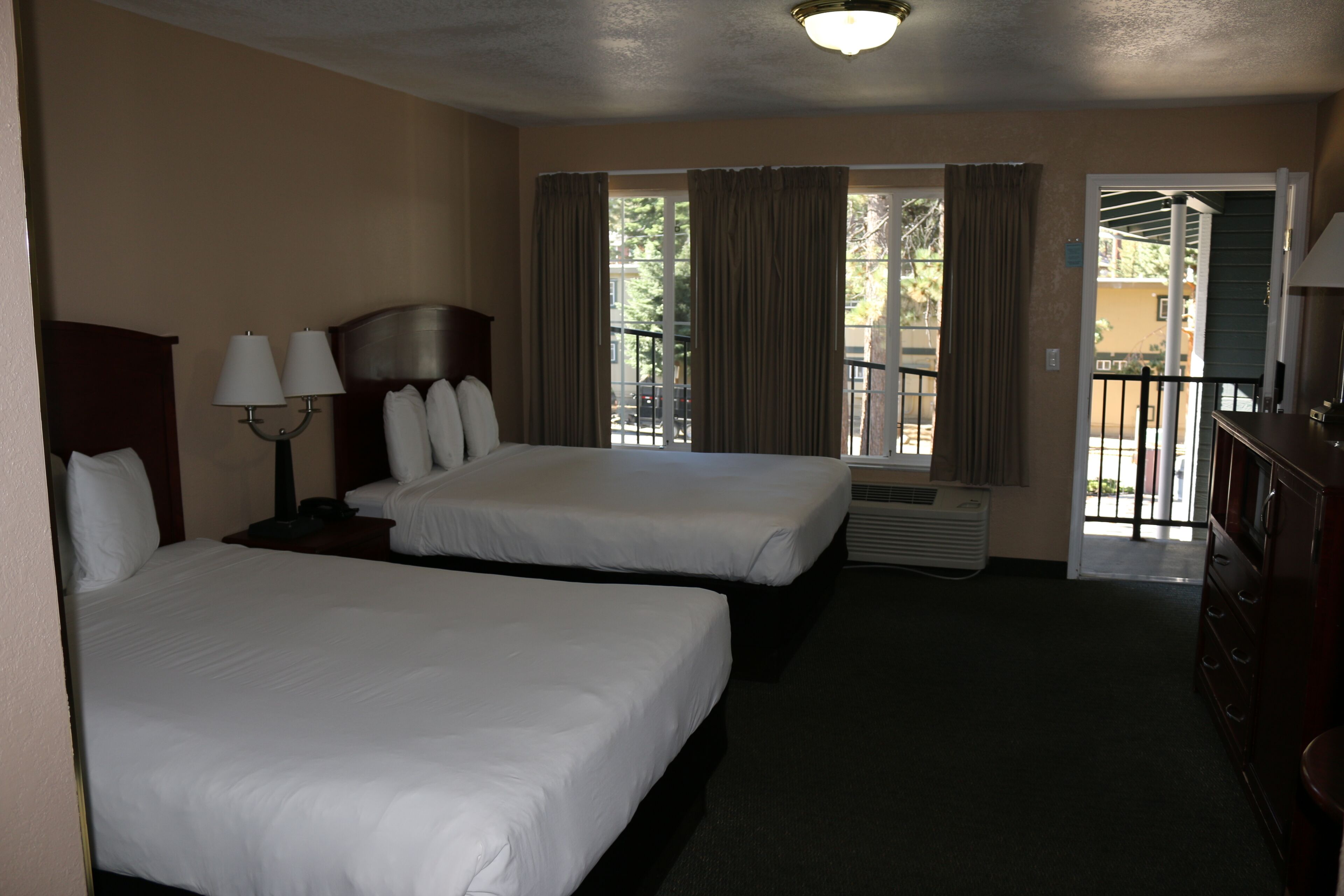 A hotel room with two beds, a nightstand with lamps, a dresser, and a view of the outside through a window with curtains.