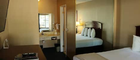 Habitación de hotel con cama, escritorio con control remoto, espejo y baño visible a través de una puerta abierta.
