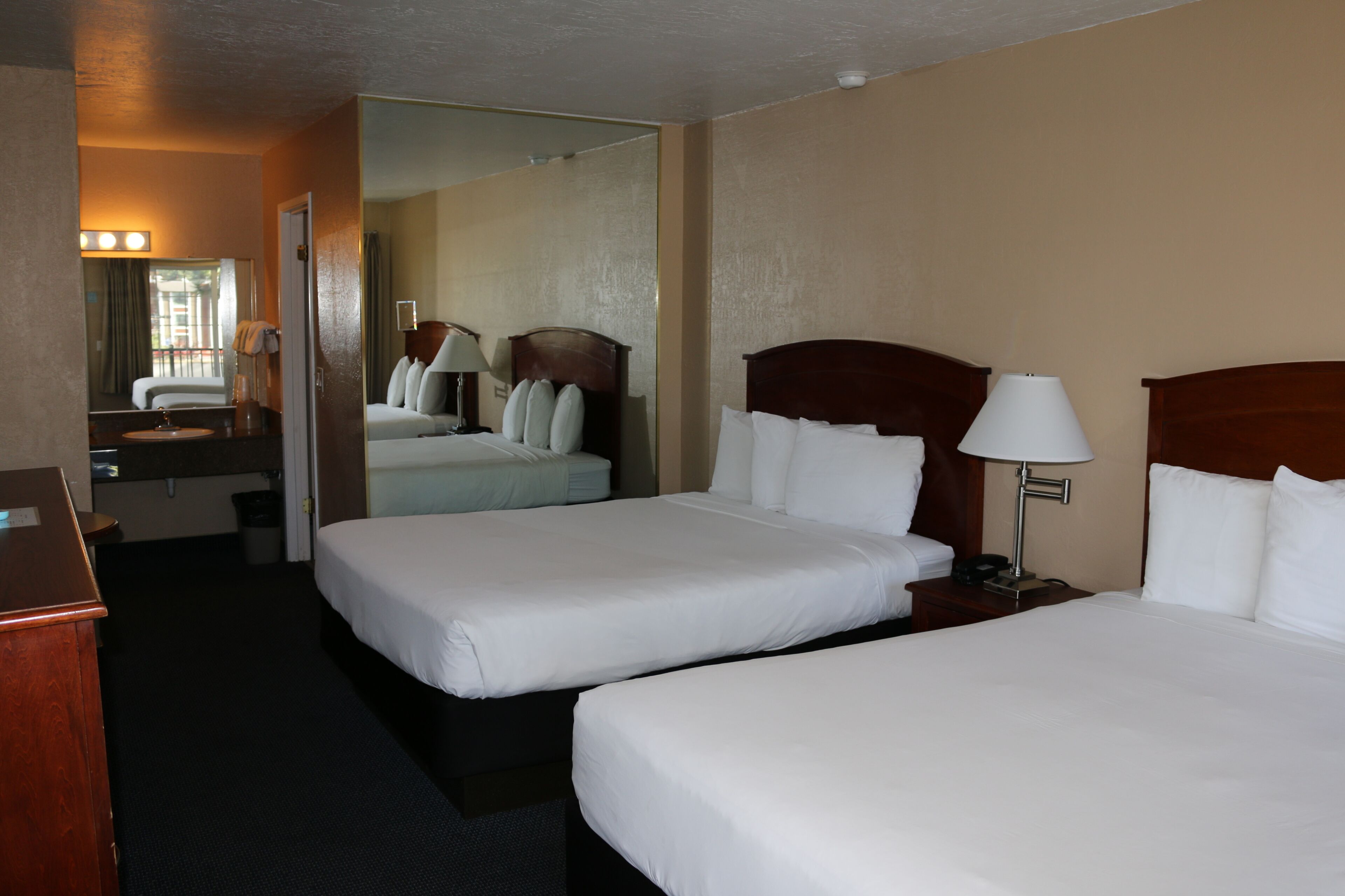 A hotel room with two single beds, a bedside table with a lamp, a mirror, and a view of the outside.