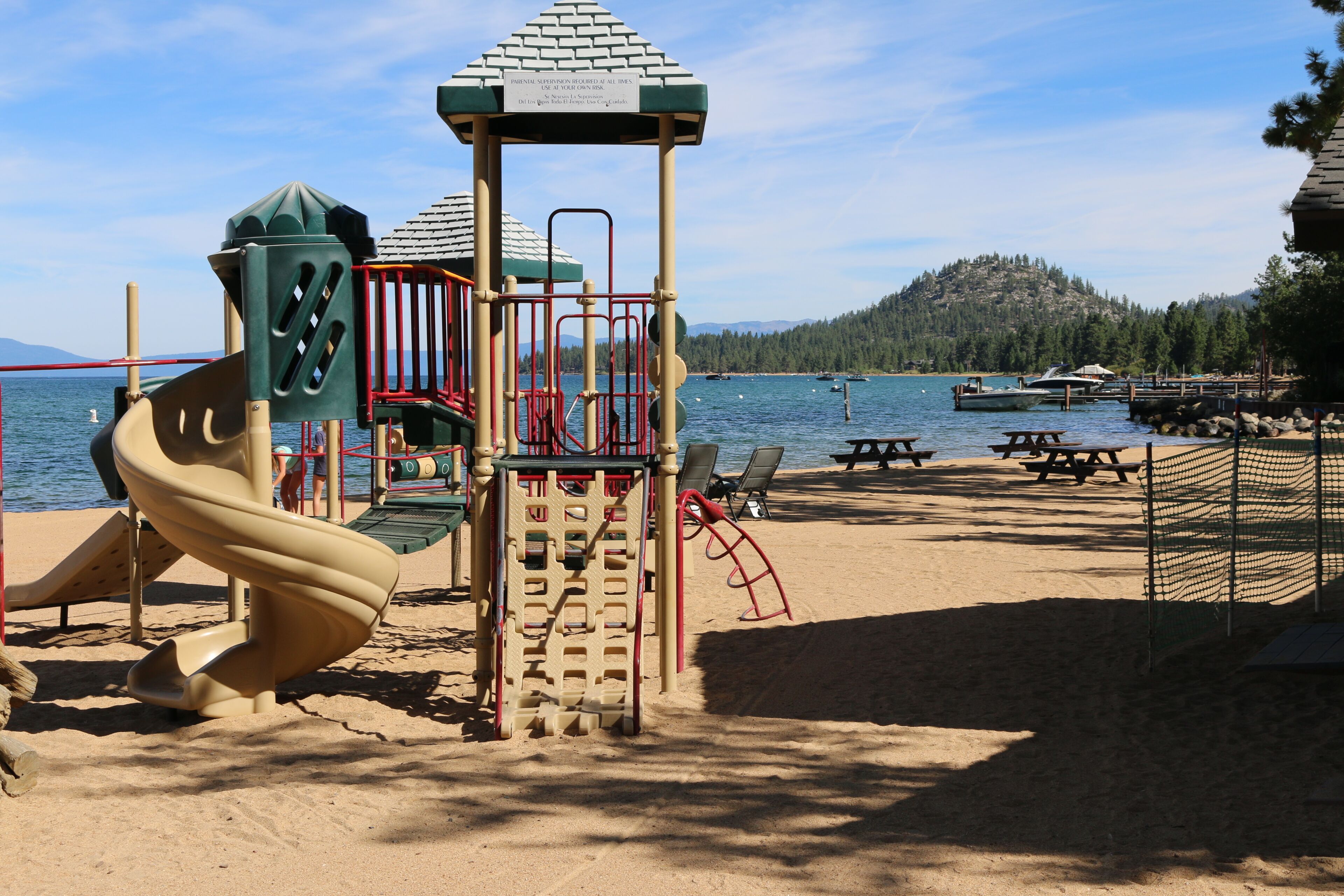 A playground with a slide and climbing structures, located near a body of water with boats and a forested hill in the background.