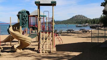 A playground with a slide and climbing structures, located near a body of water with boats and a forested hill in the background.