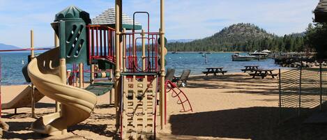 A playground with a slide and climbing structures, located near a body of water with boats and a forested hill in the background.