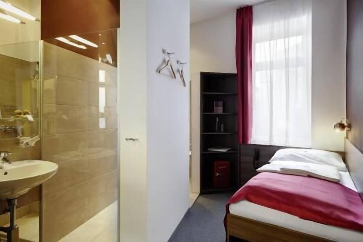 Habitación de hotel con cama, mesita de noche, espejo, armario y baño con lavabo y ducha.