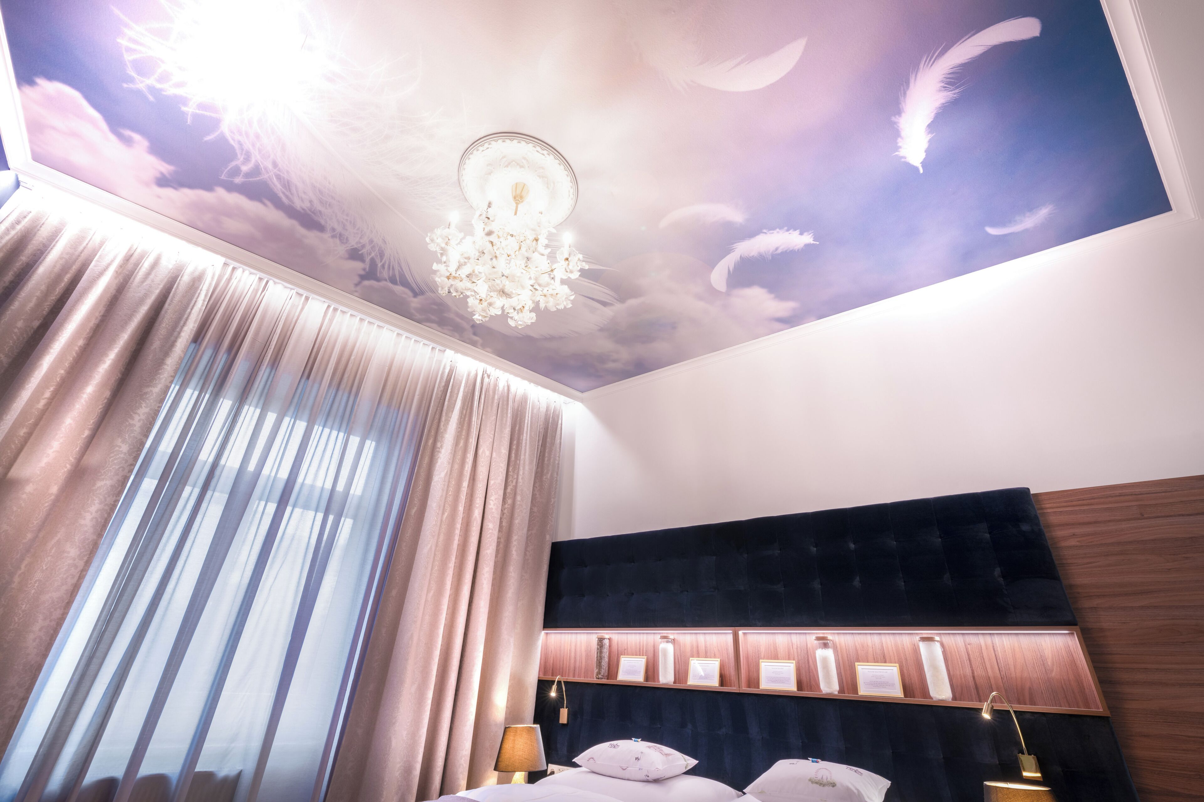 A bedroom with a large bed, a chandelier, a ceiling mural, and a window with curtains.