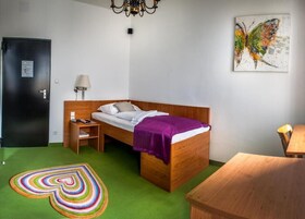 Quarto individual com cama de solteiro, cabeceira de madeira, mesa de cabeceira, luminária, tapete colorido, escrivaninha de madeira e uma pintura de borboleta na parede.