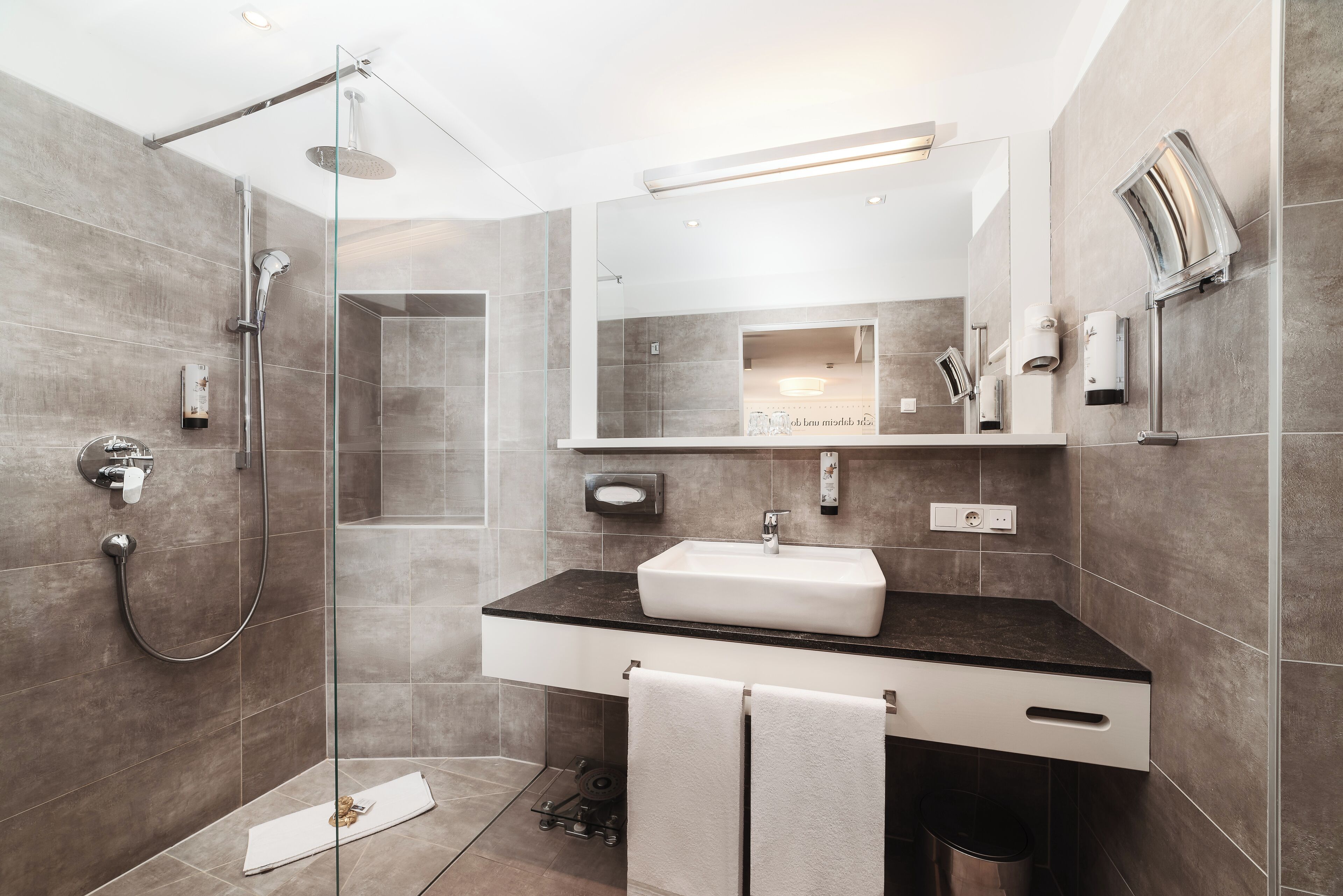 Un baño moderno con ducha envuelta en vidrio, lavamanos blanco y un espejo grande.