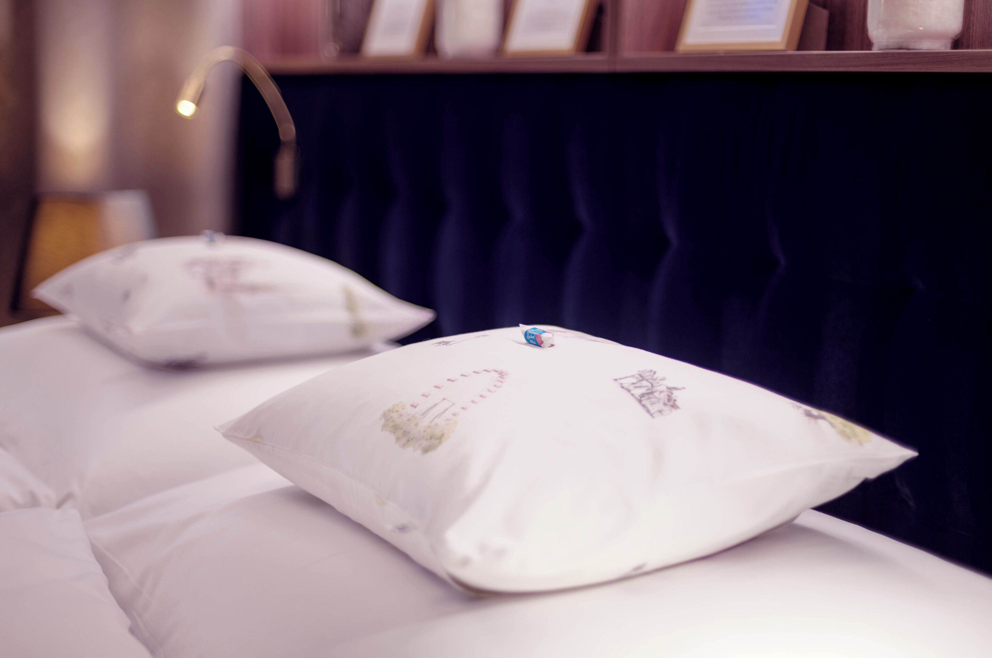 Una cama con ropa blanca y almohadas decorativas, un telón oscuro de fondo y un objeto enmarcado en la pared.