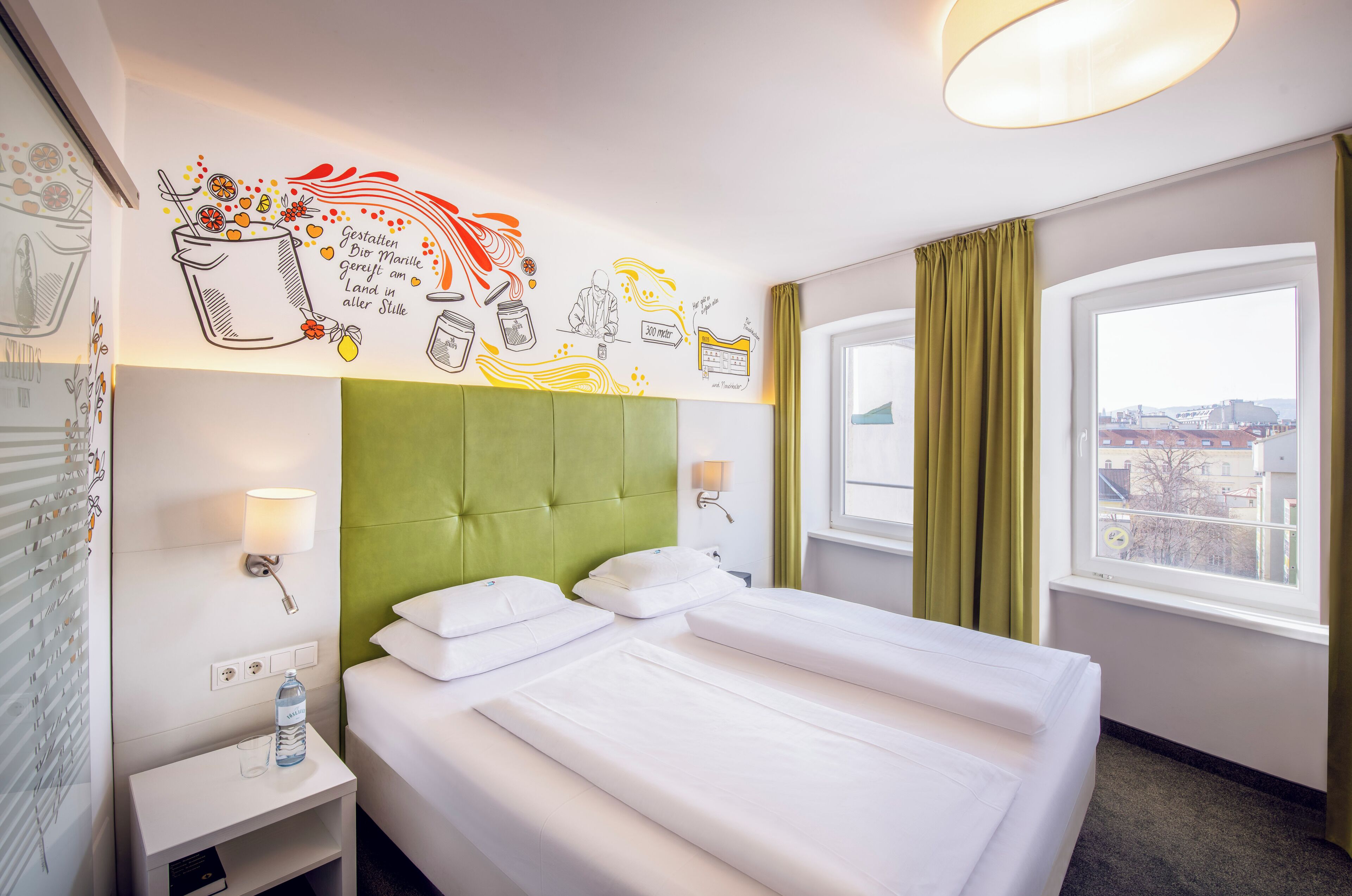 Une chambre d’hôtel avec un lit, une table de chevet, une lampe, une fenêtre avec des rideaux et un mur orné d’une fresque.