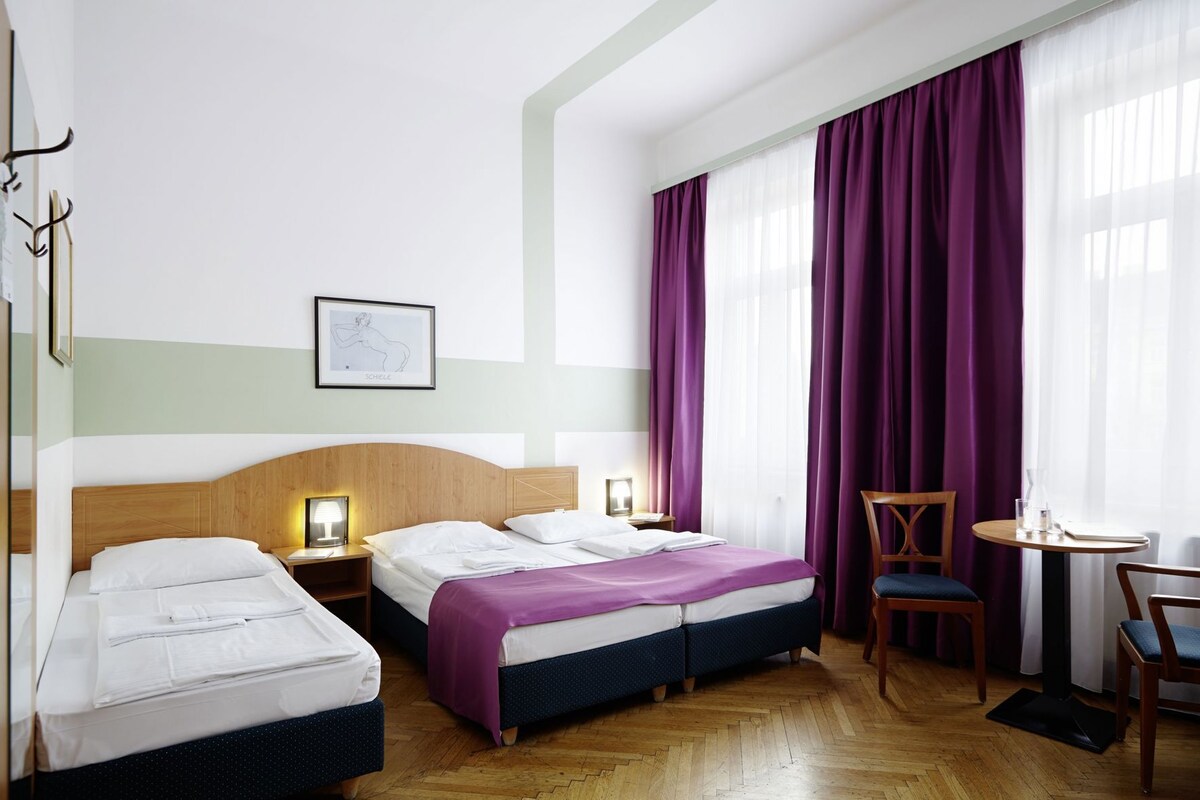 A hotel room with two beds, a wooden headboard, a small table with a chair, and a large window with purple curtains.