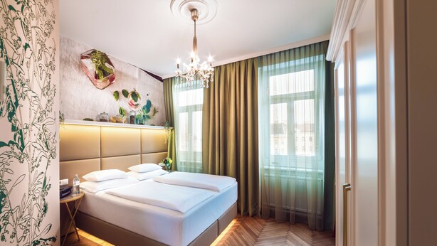 A modern hotel room with a large bed, a nightstand, a chandelier, and a view of the city through large windows. - BoutiqueHOTEL Donauwalzer (Vienna)