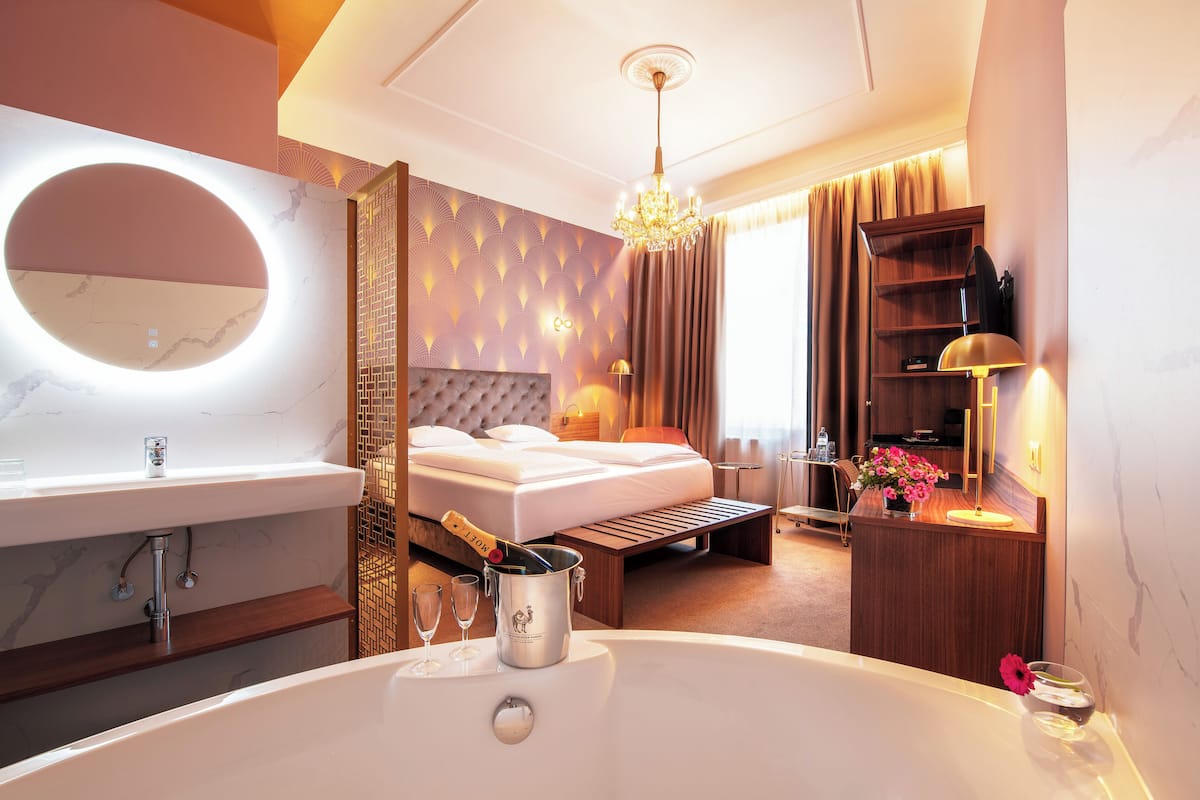Una habitación de hotel moderna con una cama grande, un área de estar separada, un minibar, un espejo redondo grande y una bañera.
