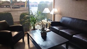 Et værelse med en lædersofa, et sofabord med en plante og udsigt til en parkeringsplads gennem et vindue.