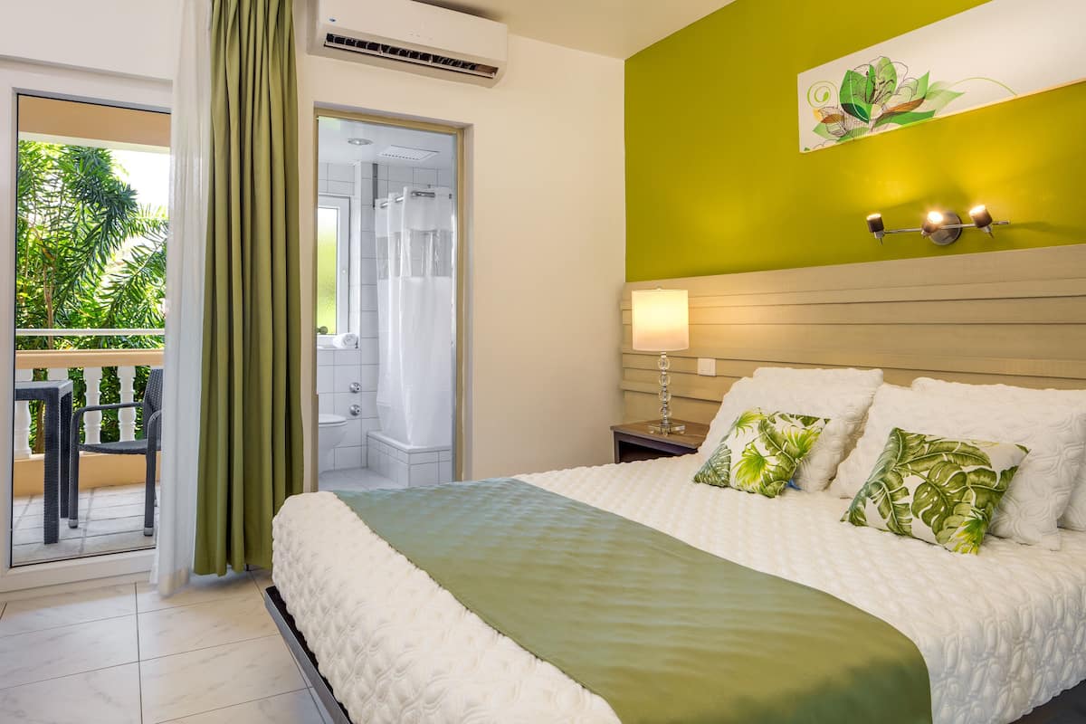 A hotel room with a bed, a green wall, a window with a view of greenery, and a bathroom visible through an open door.