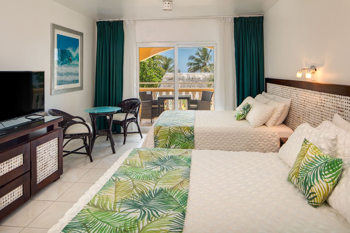 A hotel room with two beds, a TV, a desk, and a balcony with a view of palm trees.