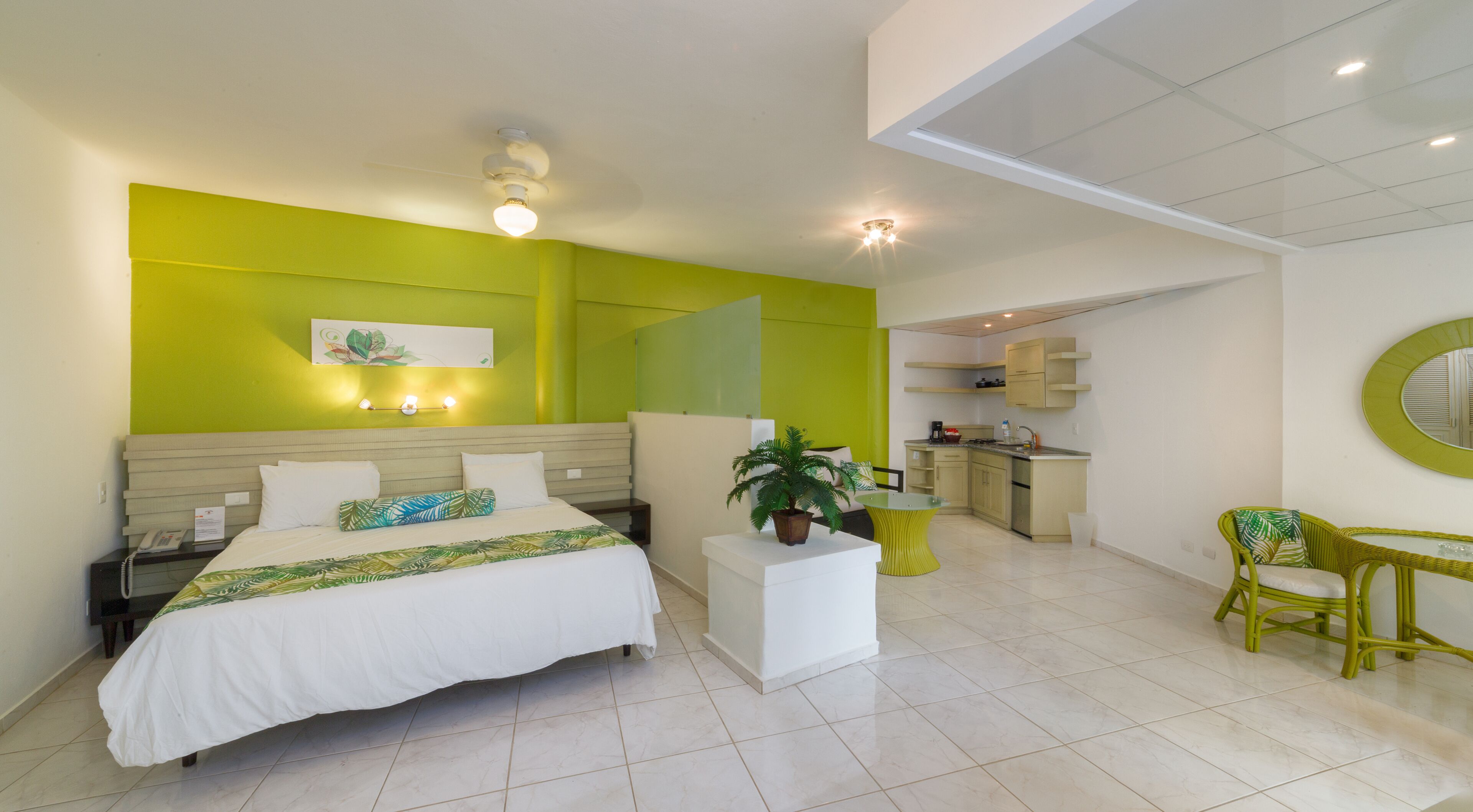 Quarto de hotel com cama, mesa de cabeceira, uma pequena planta em vaso, uma cadeira, escrivaninha e uma cozinha compacta ao fundo.
