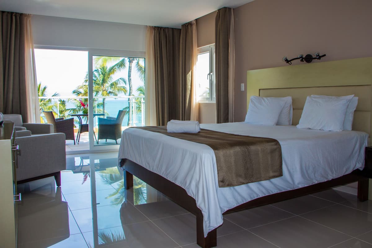 A hotel room with a large bed, a balcony with a view of palm trees and the ocean, and a chair by the window.