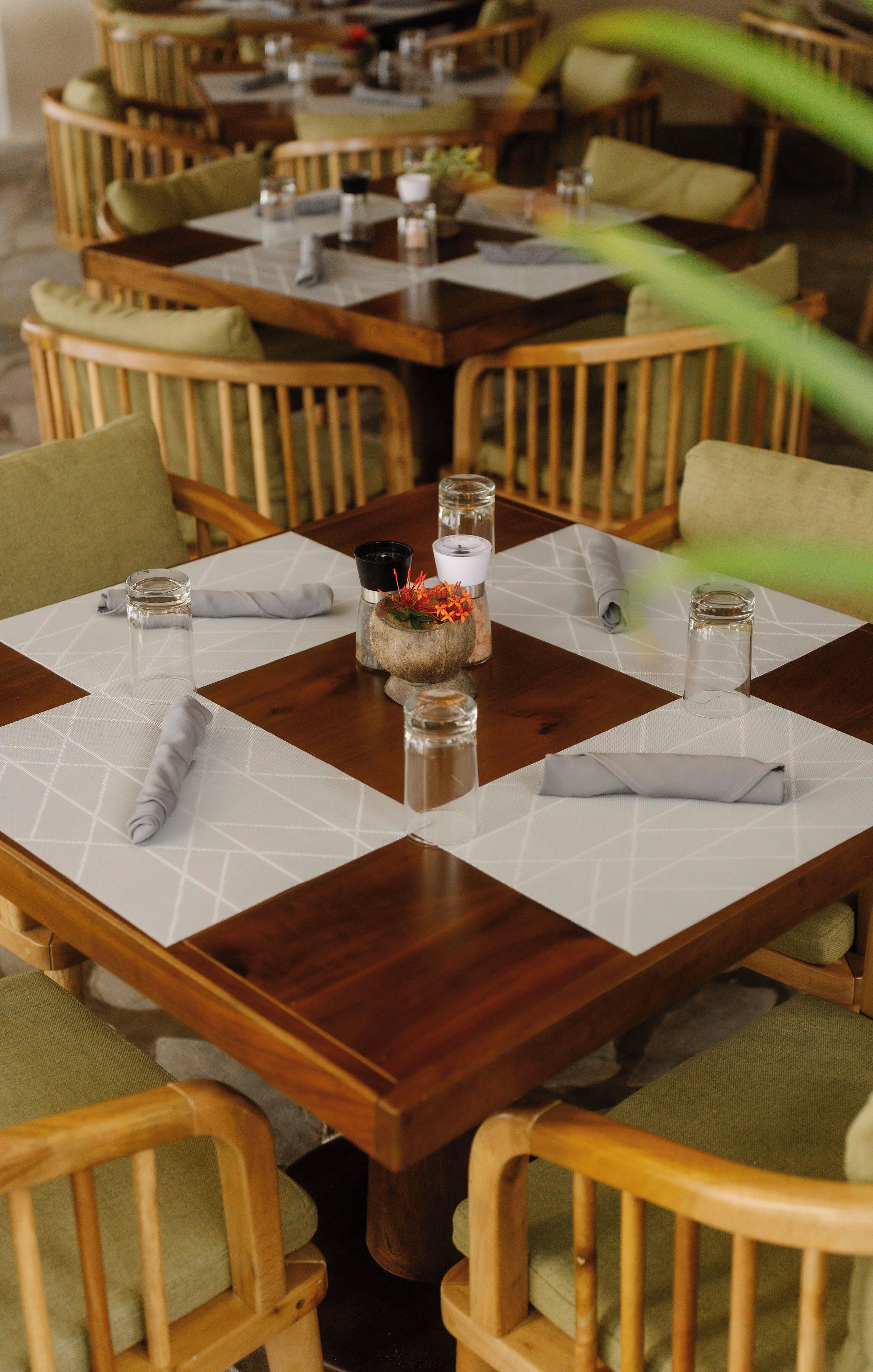 A dining area with wooden tables and chairs, each set with a white placemat, glassware, and a centerpiece.