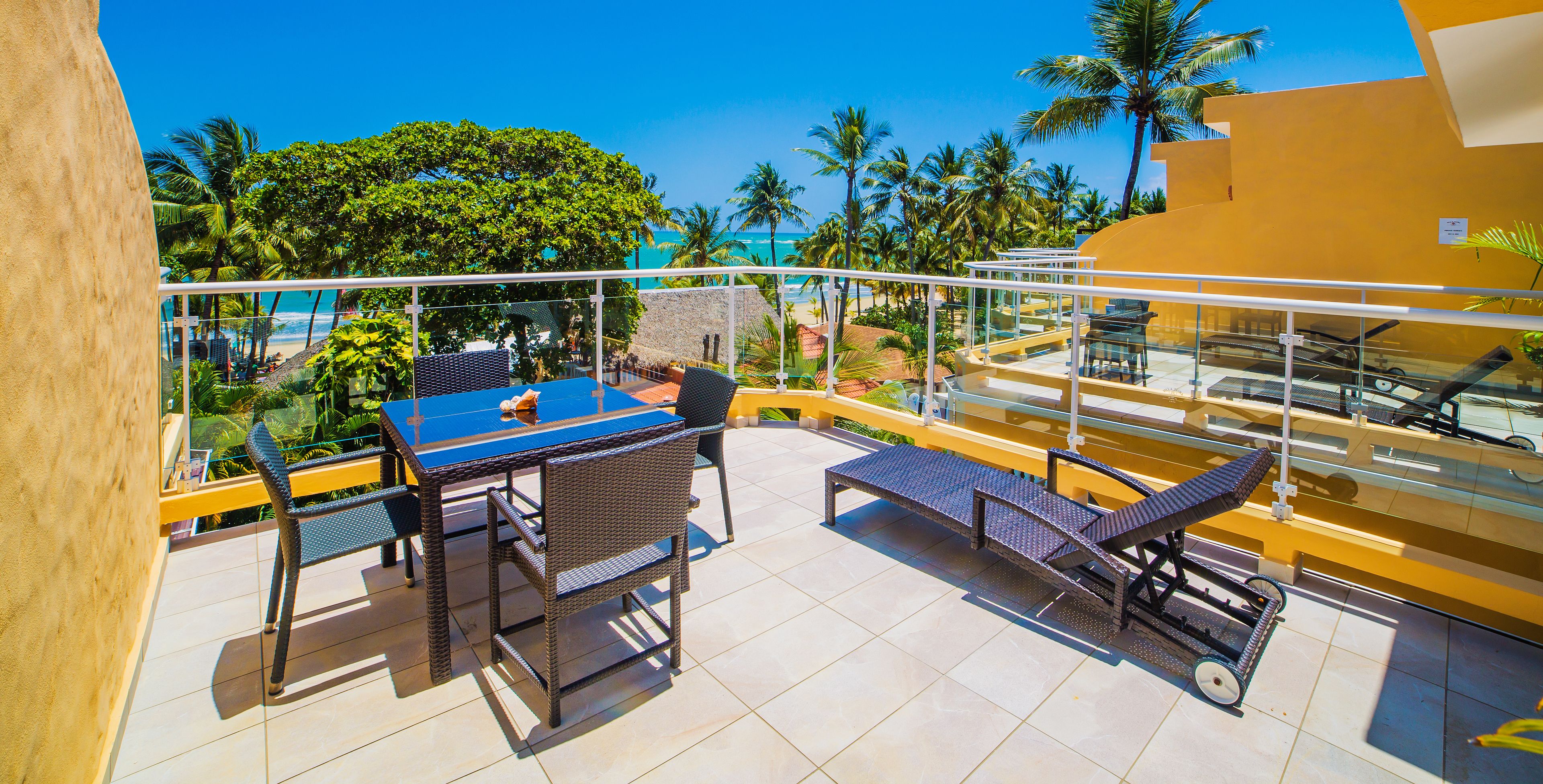 A balcony with a table and chairs, overlooking a beach and palm trees.