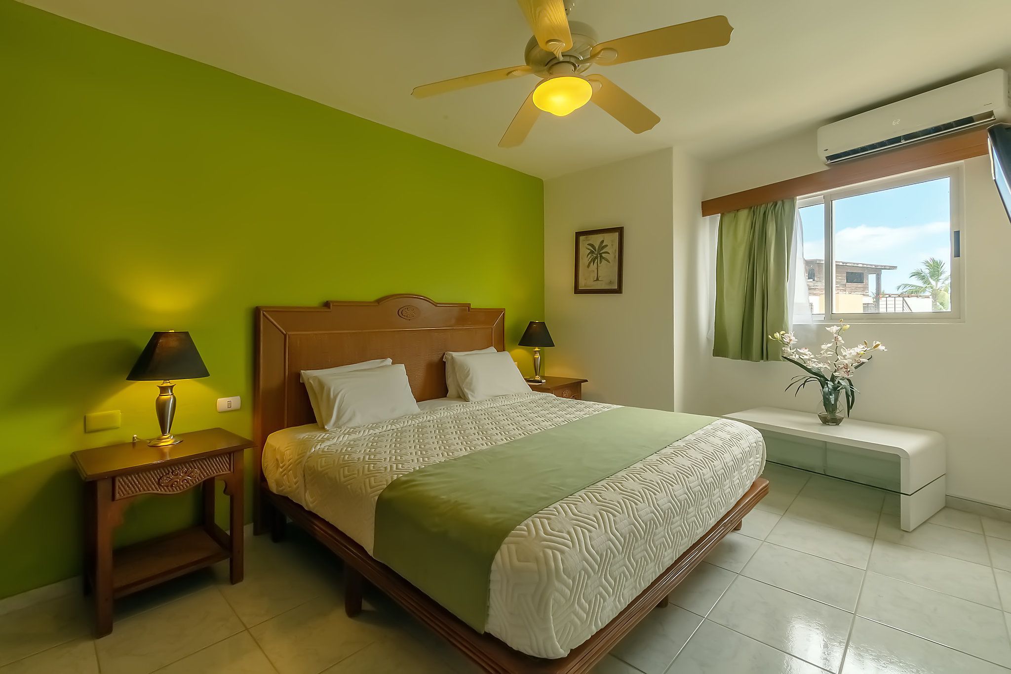 Une chambre à coucher avec un lit, deux tables de chevet, un ventilateur de plafond, une fenêtre avec des rideaux et un petit banc agrémenté d’un vase de fleurs.