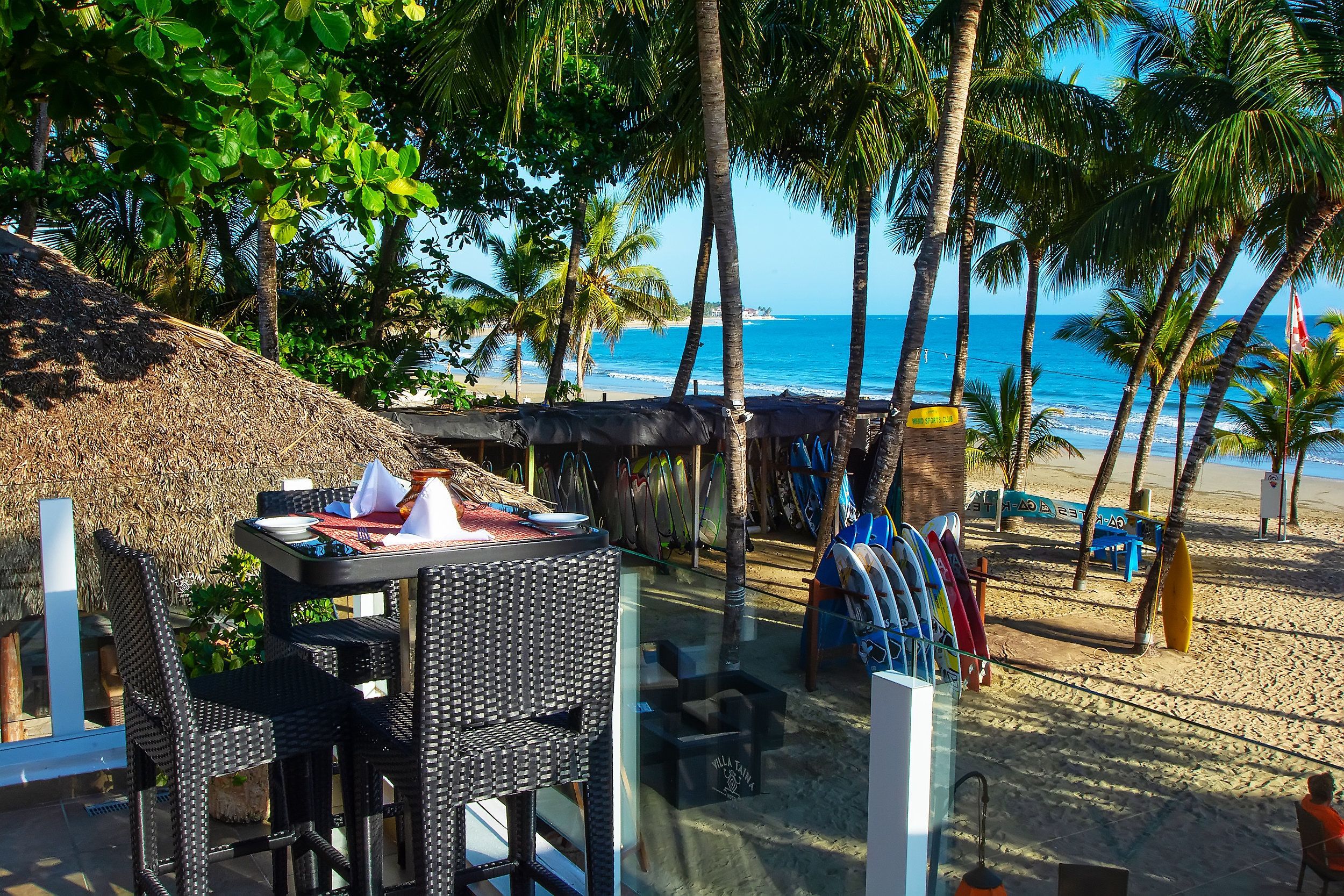 Un restaurant situé en bord de mer, avec une terrasse, un toit de chaume et des planches de surf en arrière-plan.