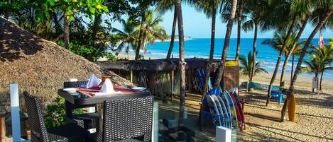 Un restaurant situé en bord de mer, avec une terrasse, un toit de chaume et des planches de surf en arrière-plan.