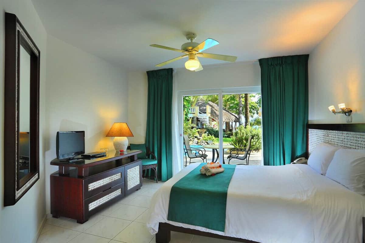 A hotel room with a bed, a TV, a desk with a lamp, a ceiling fan, and a view of the outdoors.
