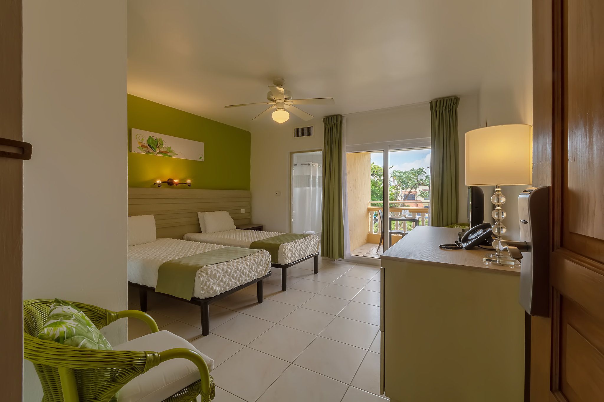 A hotel room with two beds, a desk with a phone, a chair, a ceiling fan, and a view of the outside.