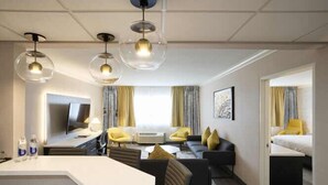Une chambre d’hôtel moderne dotée d’un espace salon central, d’un téléviseur à écran plat et d’une grande fenêtre avec des rideaux.