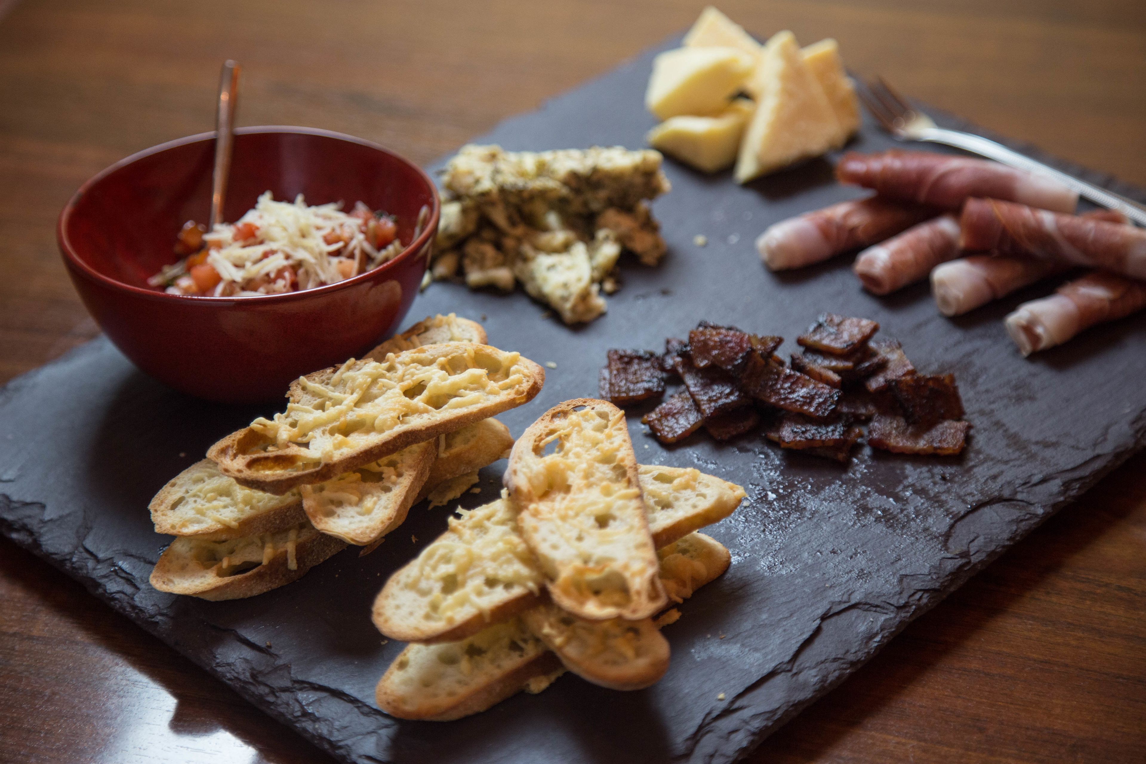 Une planche de service en ardoise, sur laquelle sont disposés divers aliments, notamment du pain, du fromage et des charcuteries.