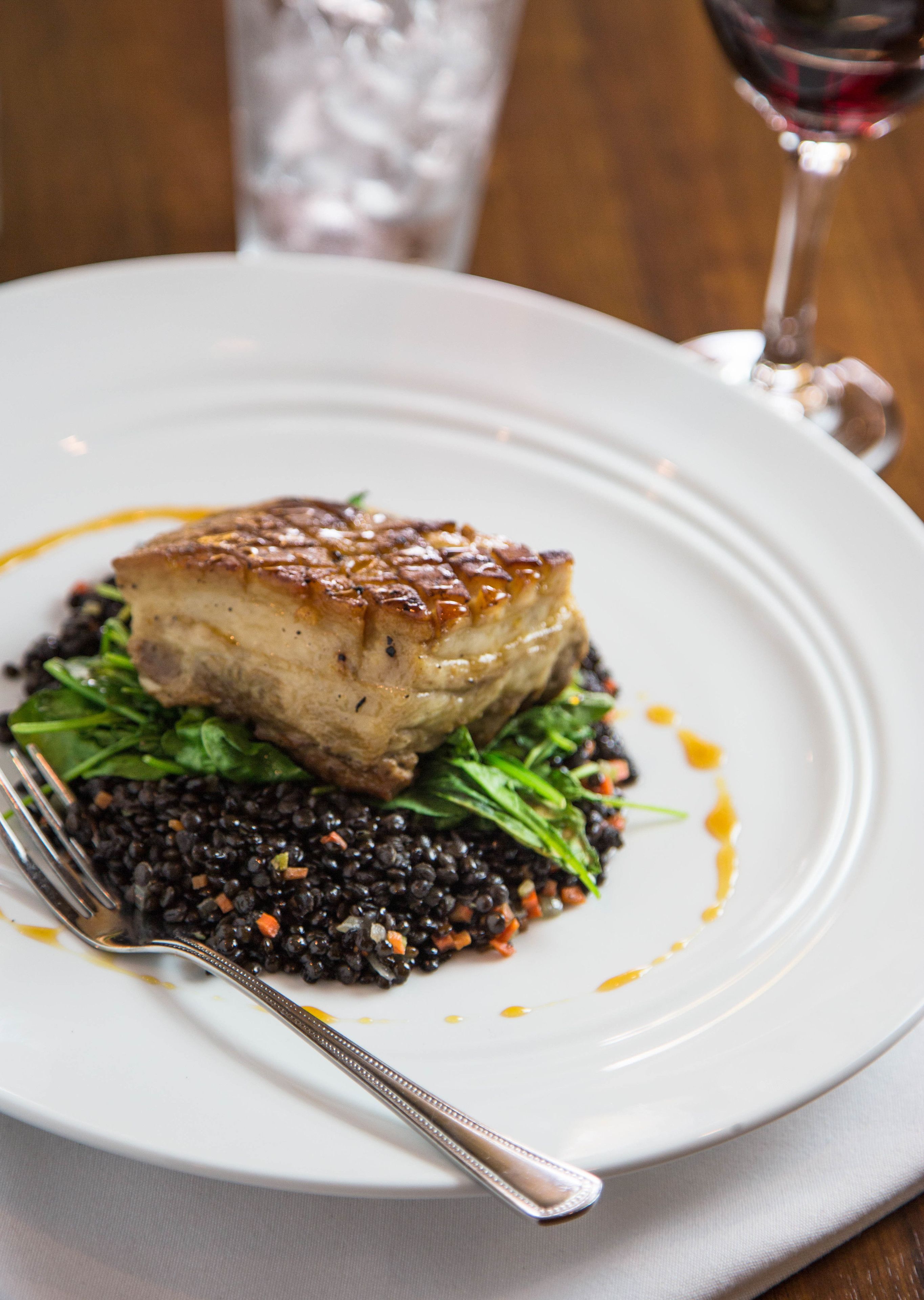 A plate with a pork chop, black lentils, and greens, accompanied by a fork and a glass of red wine.