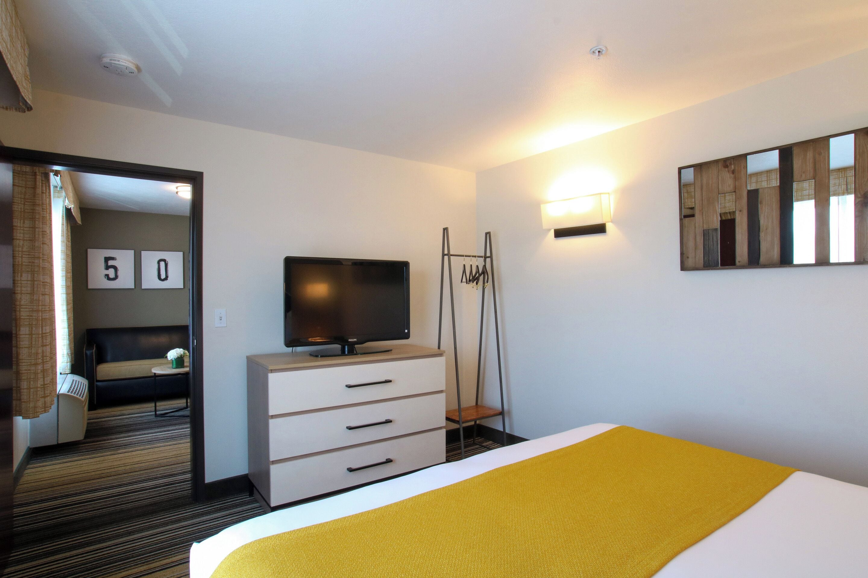 Ein Hotelzimmer mit einem Bett, einem Fernseher, einem Schreibtisch mit Schubladen, einem Spiegel und einer Tür zu einem anderen Raum.