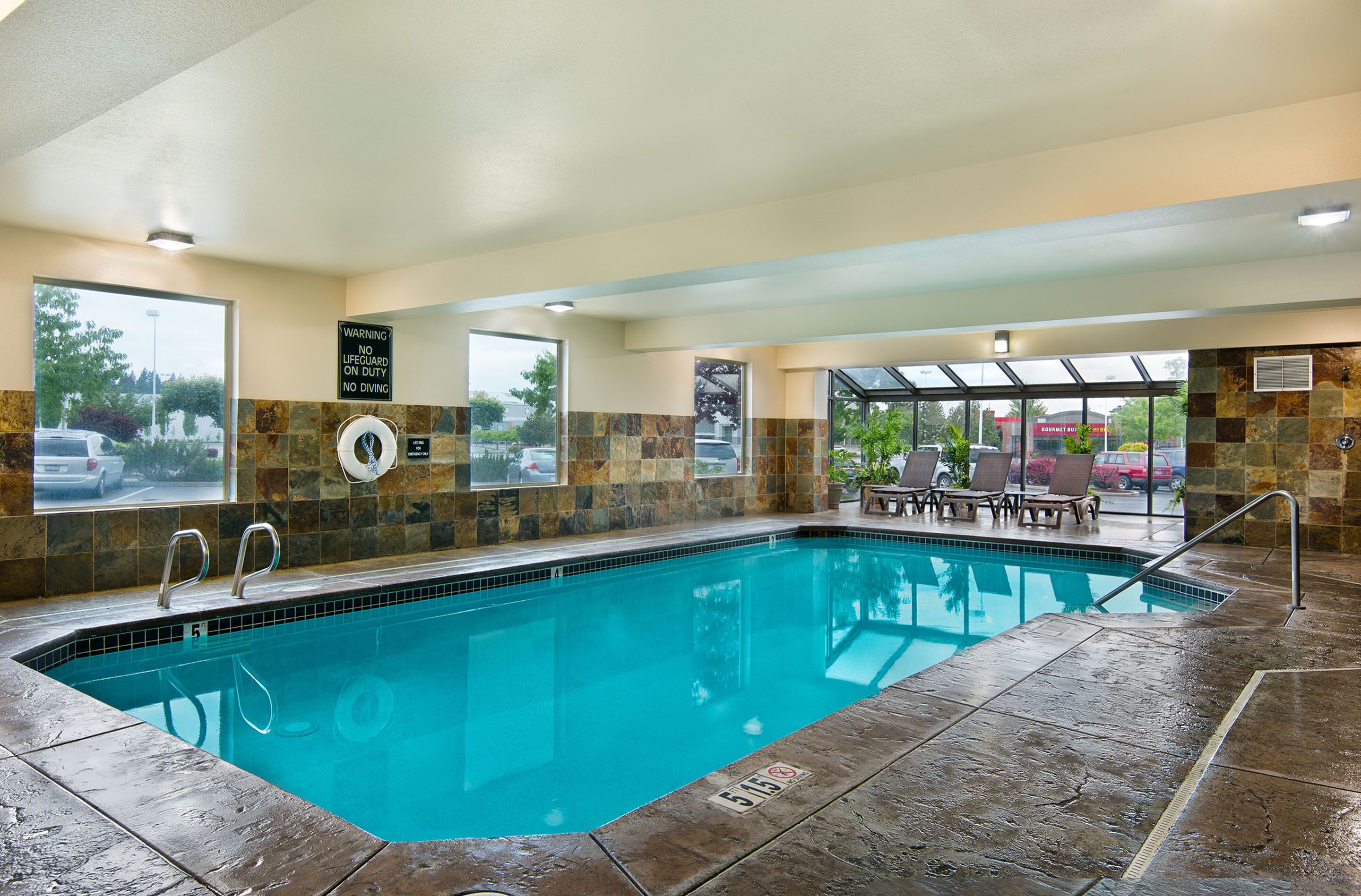 Une piscine intérieure avec un panneau indiquant la présence d’un sauveteur, des murs recouverts de carrelage en pierre et un espace détente avec des chaises et une table.