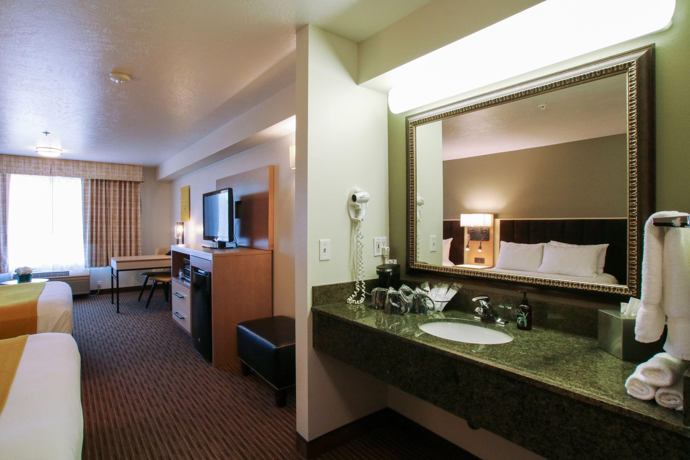 A hotel room with a bed, desk, and a large mirror above the sink.