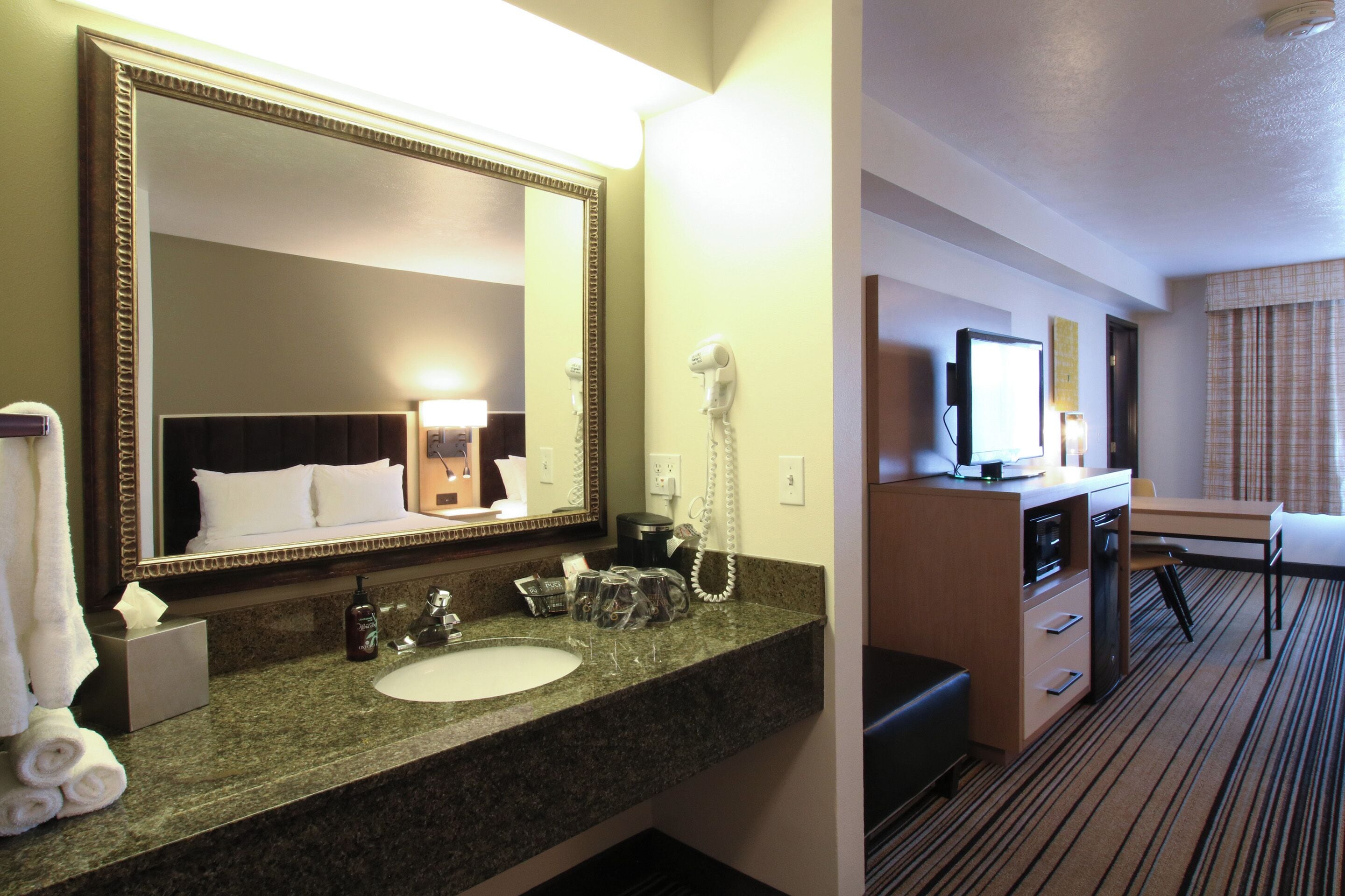 A hotel room with a large mirror, a sink, a hairdryer, a desk, a television, and a bed.