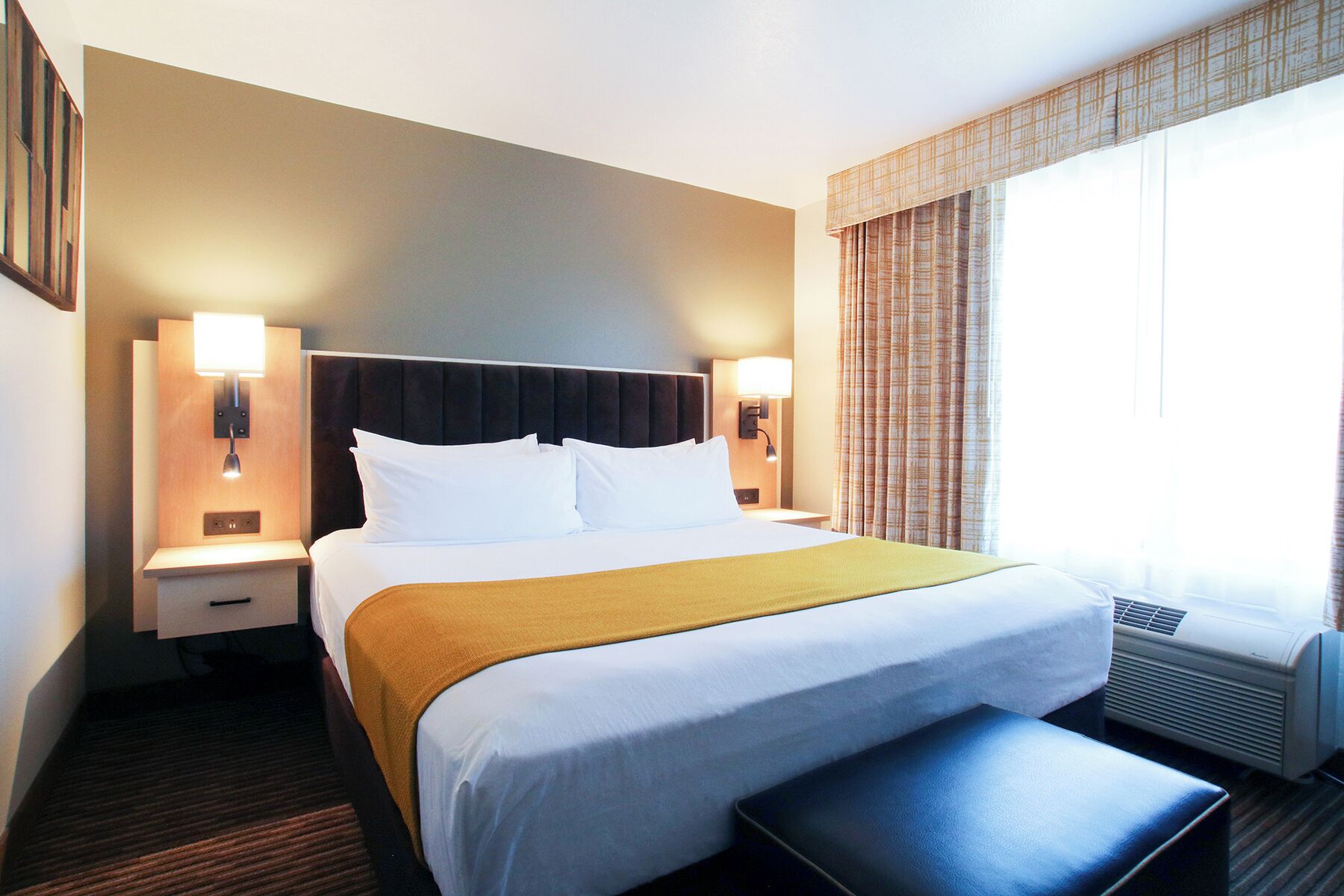 A hotel room with a bed, bedside tables, a bench, a window with curtains, and wall-mounted lights.