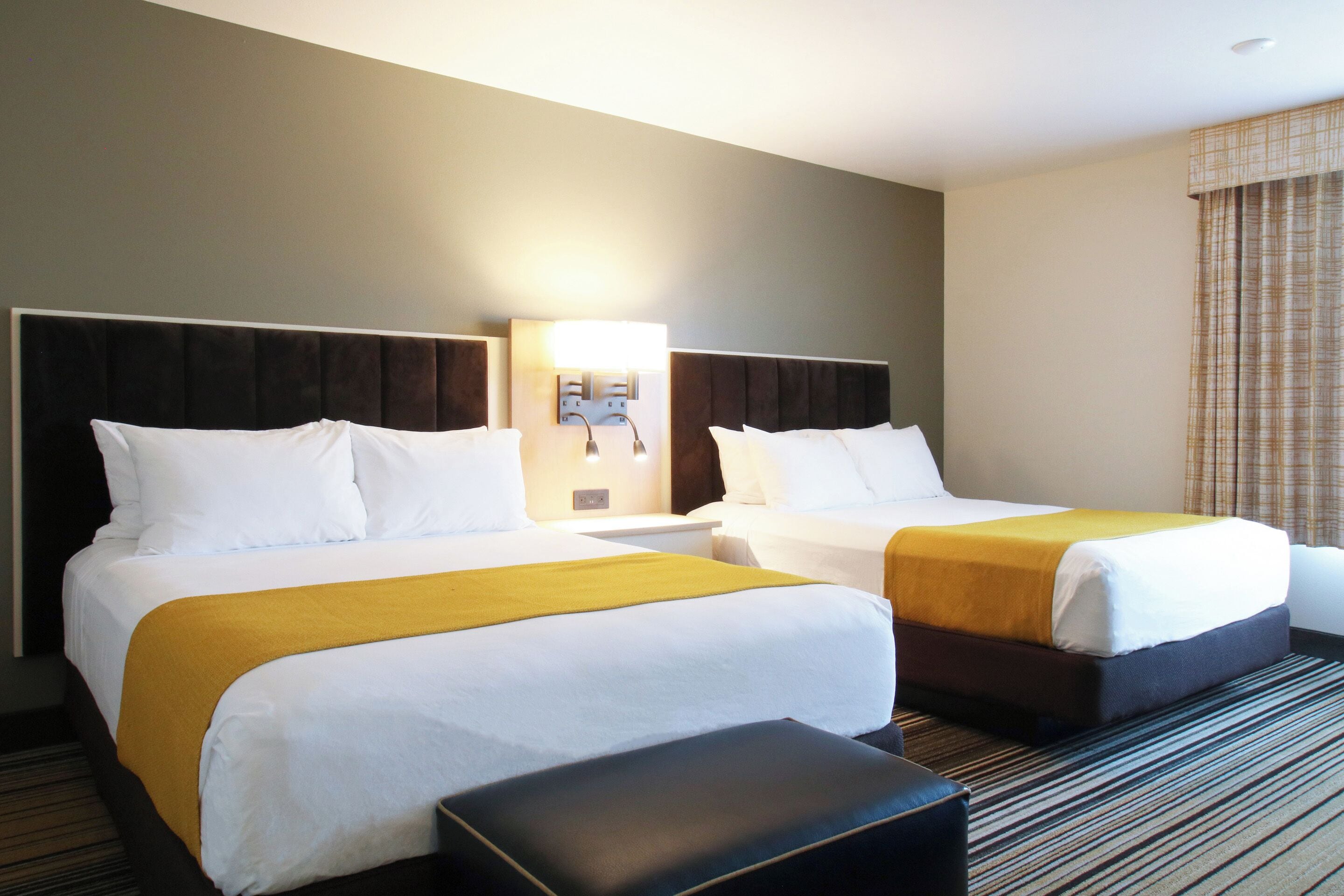 A hotel room with two beds, a nightstand, a lamp, and a window with curtains.