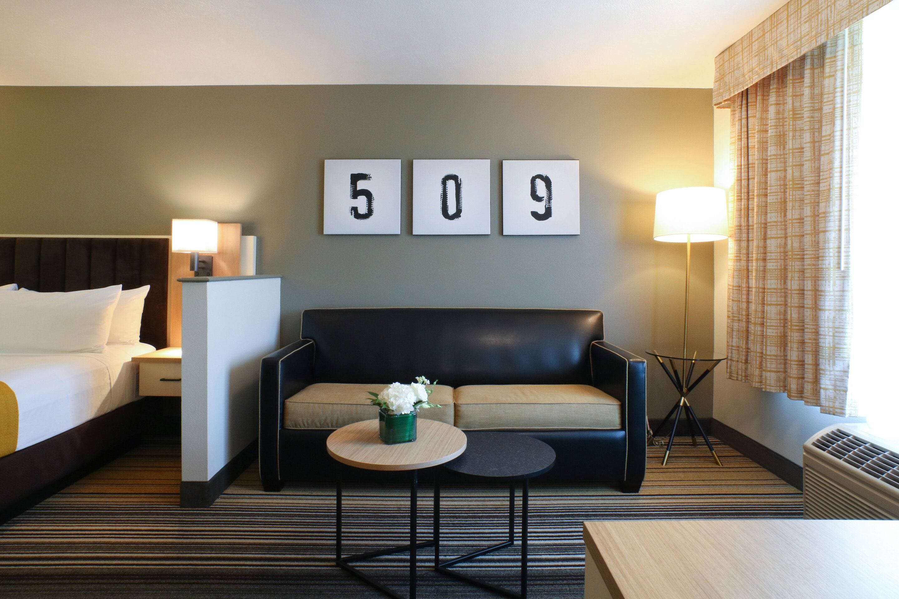 Quarto de hotel com cama, sofá, pequena mesa com um vaso de flores e um mostrador de número fixado na parede.